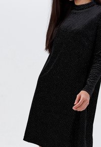 Černé šaty s dlouhými rukávy, zdobené puntíky. Vyrobené z měkké tkaniny, s vysokým výstřihem a uvolněným A-liniovým střihem.