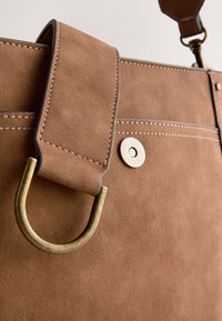 Sac à main en daim marron avec une sangle épaisse, un anneau en D en laiton, des coutures apparentes et un fermoir magnétique circulaire texturé sur fond beige.