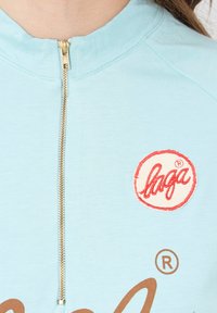 Sudadera con cierre en color azul claro, con una cremallera dorada, que presenta un logotipo circular en rojo y blanco y texto marrón en la parte delantera. Textura suave y diseño casual.