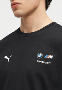 Záber zblízka osoby nosiacej čierne tričko s logom Puma na ľavej strane a logami BMW Motorsport na pravej strane hrudníka.