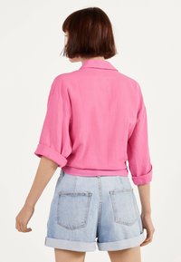 Osoba z krótkimi brązowymi włosami nosi różową koszulę z długim rękawem oraz podwiniętymi mankietami, a także jasnoniebieskie szorty dżinsowe, stojąc na białym tle.