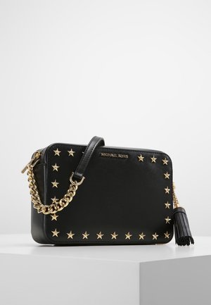 Sac bandoulière rectangulaire noir Michael Kors avec clous étoiles dorés, bandoulière chaîne et détail pompon en cuir sur surface blanche.