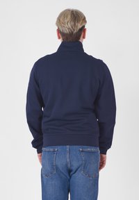 Giacca a zip blu navy con colletto alto, polsini e orlo a costine, realizzata in un tessuto liscio. Indossata con jeans in denim azzurro.
