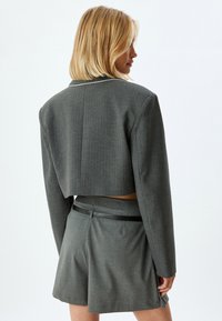 Harmaa lyhyt bleiseri, jossa on jämäkkä muotoilu, varustettuna sakara-kauluksella ja hienovaraisella houndstooth-kuviolla, yhdistettynä vastaaviin räätälöityihin shortseihin.