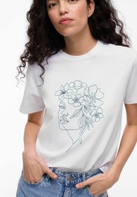 Hvid t-shirt med en linje-tegnet blomsterdesign, der rammer et ansigt. T-shirten er lavet af bomuldsstof, har korte ærmer og en afslappet pasform.