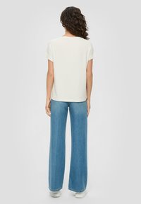 Helles beige Kurzarmoberteil kombiniert mit hochtaillierten, weit geschnittenen blauen Jeans. Glatter Stoff mit einer lockeren Passform, von hinten gezeigt.