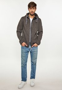 Sivi, zip-up dukserica s podesivim vrpcama, s detaljima od krpa, u kombinaciji sa svijetloplavi trapericama i bijelim tenisicama.
