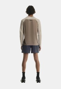 Langärmliges, hellbeiges Sportoberteil mit einem braunen strukturierten Rücken, kombiniert mit navyblauen Shorts und schwarzen Schuhen. Verfügt über ein schlankes Design und eng anliegende Ärmel.