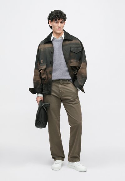 Μαύρο μπουφάν με καφέ κλίση, γκρι πουλόβερ, μπεζ παντελόνι και λευκά αθλητικά παπούτσια. Κρατάει μια μαύρη τσάντα. Απλό φόντο, ουδέτεροι τόνοι.