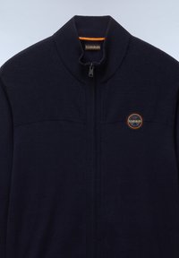 Marinblå zip-jacka med en texturerad övre panel, slät nedre del och en logotyp på vänster bröst. Hög krage och dragkedjestängning.