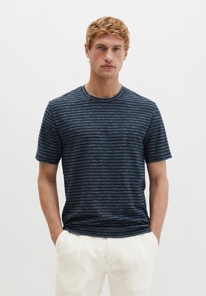 Man draagt een marineblauw-zwart gestreept T-shirt met korte mouwen en witte broek, staat met handen in de zakken tegen een effen lichte achtergrond.