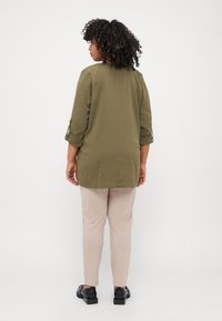 Tunic verde oliv cu mâneci trei sferturi, material texturat, cu fante laterale, asortată cu pantaloni bej slim-fit și pantofi negri.