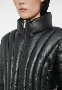 Svart puffjakke med høy krage, med en blank tekstur, vertikal quiltet design og gullglidelåsdetalj.