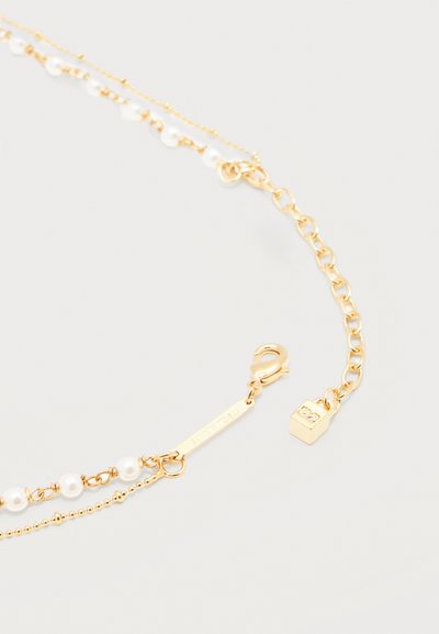 Collier superposé en ton or avec petites perles blanches, fermoir mousqueton, chaîne rallonge et pendentif en forme de cube sur un fond neutre.