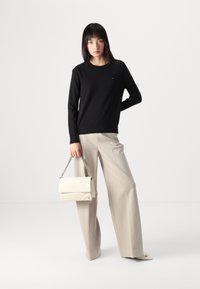 Maglione nero a maglia, pantaloni beige a gamba larga, tiene in mano una borsa in pelle crema con tracolla a catena. Design semplice e colori neutri.