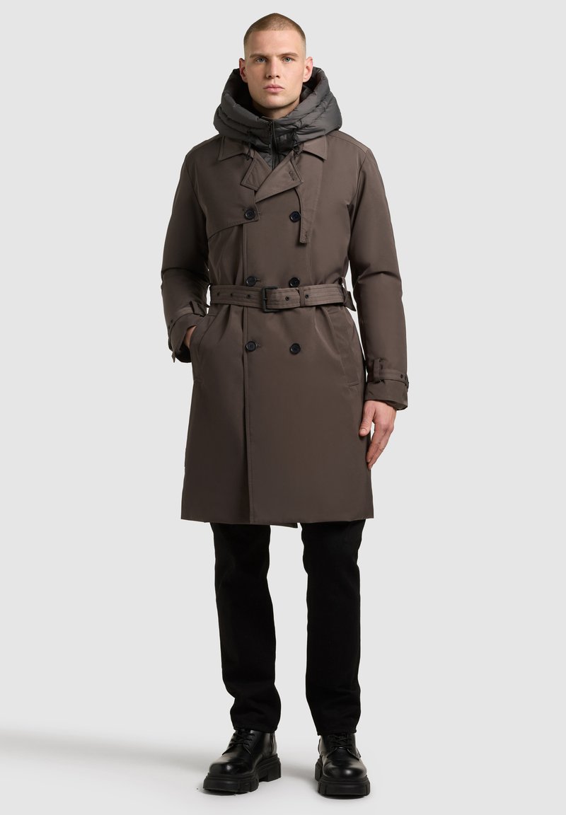 Brown trench coat with a detachable gray hood, double-breasted design, belted waist, and side pockets, styled with black pants and shoes.