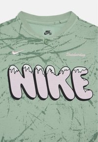 Zelená košile s krátkým rukávem, která má strukturovaný vzor, zapínací límec, velké černé a růžové logo "NIKE" a nápis "Skateboarding" ve světle růžové barvě.