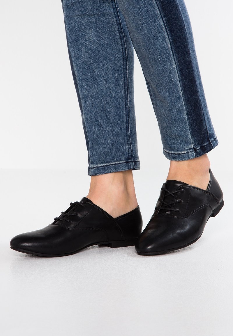 Chaussures en cuir noir à profil bas, bout pointu et design à lacets, associées à un jean bleu avec un panneau latéral contrastant.