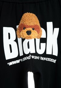 Pantalones de chándal negros con grandes letras blancas y un aplique de cara de perro marrón de felpa y textura en el frente. Material de mezcla de algodón.