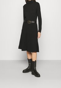 Robe noire en tricot côtelé à manches longues, col haut et taille définie, agrémentée d'une ceinture en cuir noir et de bottes noires épaisses.