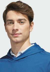 Modrá mikina s kapucí z texturované látky, s kapucí a vrstveným límcem přes světle modrou košili. Detailní portrét mužského modelu.