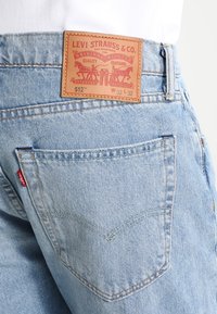 Ljusblå denimjeans med en brun läderlapp vid midjan som har präglad text. Inkluderar en bakficka med en V-format sömdetalj.