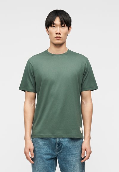 Άνδρας που φοράει έναν απλό πράσινο κοντομάνικο μπλουζάκι και μπλε τζιν, στέκεται μπροστά σε ουδέτερο φόντο, κοιτάζοντας προς τα εμπρός.