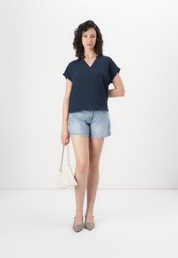 Dunkelblaues kurzärmliges Oberteil mit V-Ausschnitt, kombiniert mit hellen Jeansshorts. Das Model hält eine kleine weiße Handtasche und trägt beige spitze High Heels.