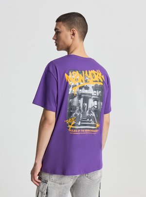 Giovane uomo con t-shirt viola con graffiti giallo "New York" e foto in bianco e nero di persone su un gradino, visto di profilo.