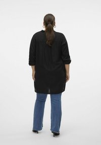 Sort, langærmet tunika med slids i siden og knaplukning ved ærmerne, parret med blå flagrende jeans og sorte hæle, set fra ryggen.