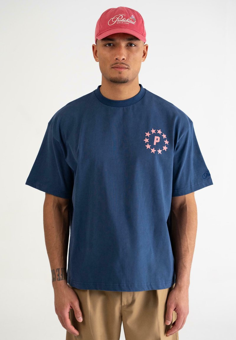 Mørkeblå bomulds T-shirt med korte ærmer, prydet med et pink stjerne-cirkel design og "P" logo på venstre bryst. Sættet er kombineret med khaki bukser.