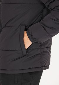 Sort pufferjakke med struktureret overflade, elastiske ærmer og en sidelomme. En quiltet design tilføjer dimension og isolering.