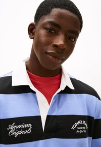 Polo a righe in azzurro, nero e bianco; presenta un colletto, loghi ricamati ed è indossata sopra una maglietta rossa.