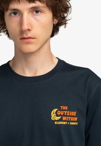 Dark blue cotton t-shirt featuring a graphic print in orange and yellow, displaying the text "THE OUTSIDE WITHIN ELEMENT & TIMBER."