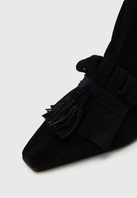 Μαύρο σουέτ μποτάκι αστραγάλου με μυτερή μύτη, διακοσμητικές φούντες και υφή. Η σόλα είναι κομψή και μίνιμαλ.