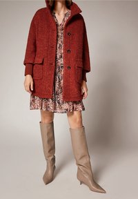 Un manteau en peluche de couleur rouille, avec un col haut, de grandes poches et des boutons noirs, superposé sur une robe à motifs. Des bottes pointues couleur beige, arrivant au genou.