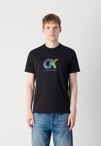 Juodas medvilnės marškinėliai su daugiamačių spalvų "OK" logotipu ir "Calvin Klein" tekstu priekyje. Modelis dėvi šviesiai mėlynas džinsines kelnes.