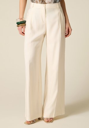 Femme portant un pantalon crème taille haute à jambes larges, un haut transparent à motifs, des bracelets épais et des sandales à talons ouvertes.