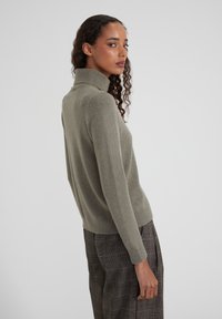 Frau mit lockigem Haar, die einen olivgrünen Rollkragenpullover und graue Karohosen trägt, seitlich vor einem einfachen Hintergrund stehend.