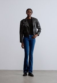 Jachetă neagră de motociclist din piele, top negru mulat, blugi evazați albaștri și pantofi negri. Modelul stă pe un fundal simplu, deschis la culoare.