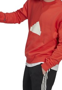 Person i röd sweatshirt med en vit trappformad logotyp och svarta byxor med vita sidostraper, hand i fickan.