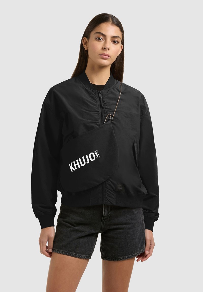 Jeune femme aux longs cheveux bruns portant une veste bomber noire, un short en jean noir et un sac à épaule noir de la marque KHUJO.