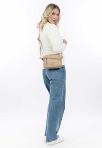 Tan crossbody bag with a zippered front pocket, rectangular shape, smooth leather texture, and adjustable strap, worn against a light sweater.