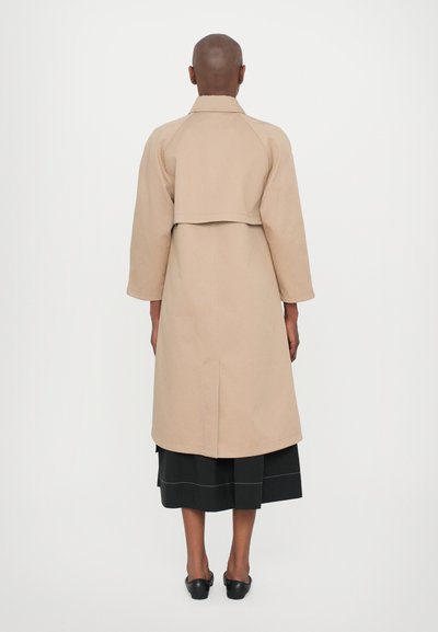 Cappotto beige lungo con una vestibilità sartoriale, maniche raglan e un design accorciato. Presenta una cucitura visibile sul retro e arriva sotto il ginocchio.