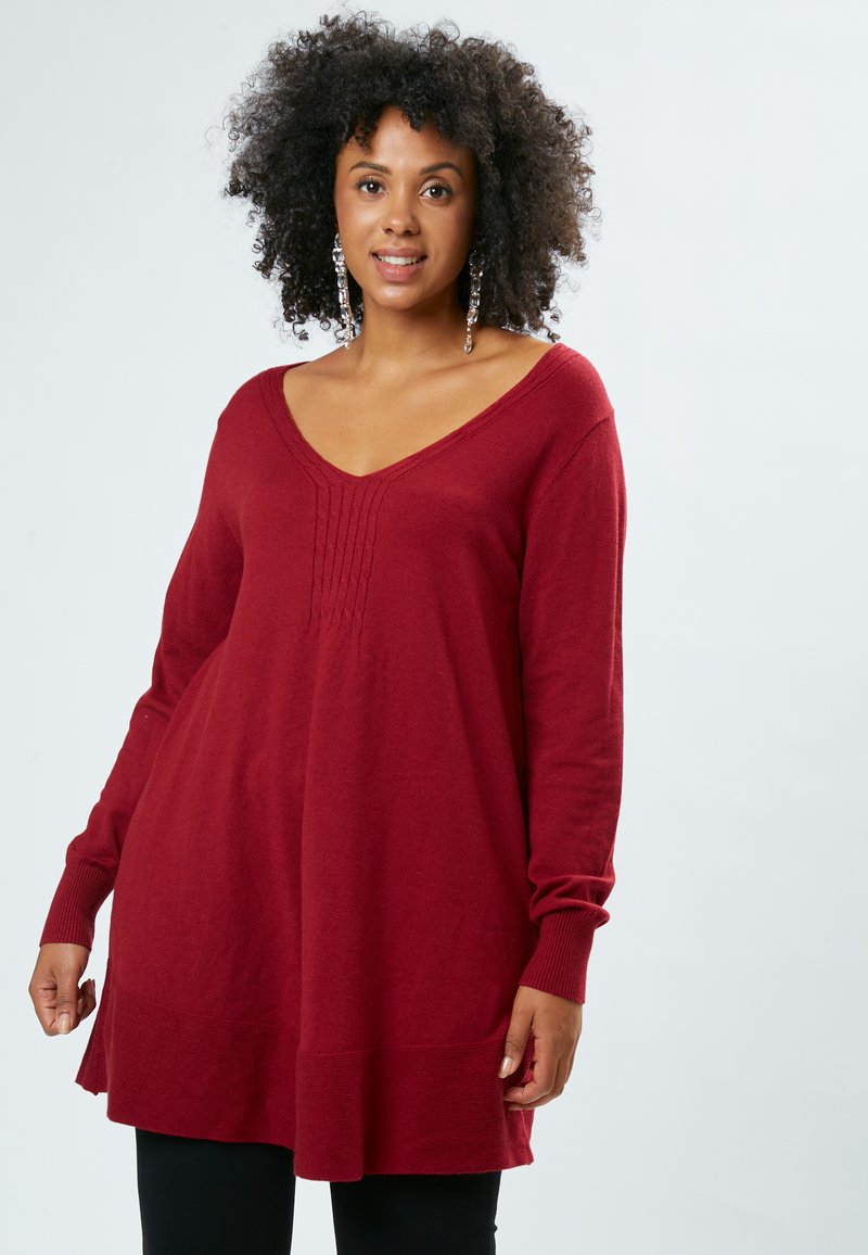 Roter Strickpullover mit V-Ausschnitt, langen Ärmeln und subtilen vertikalen Nähten auf der Vorderseite. Weiche Textur, verlängerte Passform.