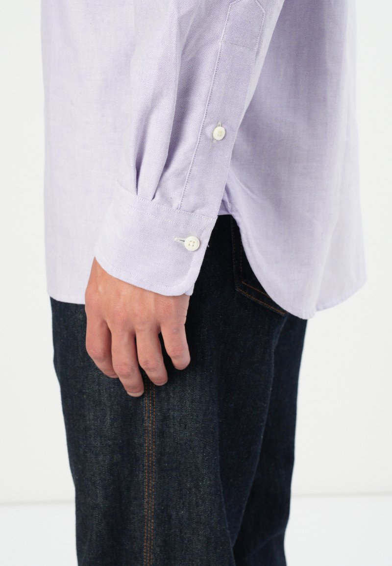 Mano descansando a un lado, vistiendo una camisa de botones de color púrpura claro y jeans oscuros con costuras visibles en el bolsillo.
