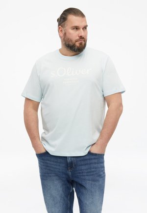 Mann mit Bart und zurückgebundenem Haar, der ein hellblaues s.Oliver T-Shirt und blaue Jeans trägt, steht mit den Händen in den Taschen und schaut zur Seite.