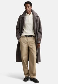 Lang brun ternet frakke med brede revers, cremefarvet kabelstrikket sweater, beige bukser og sorte sko. Bemærkelsesværdig tekstur og klassisk design.