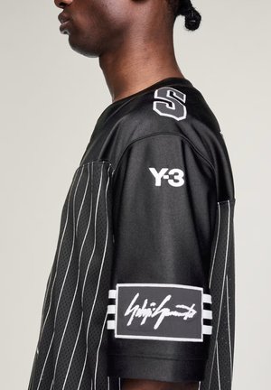 Person iført en sort sportstrøje med hvide tynde striber, "Y-3" tekst på skulderen og en stor hvid signatur-patch på ærmet.