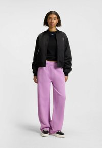 Svart bomberjacka, kort design med snapknappar, parat med ljuslila vida byxor i sweatpants-stil. Slätt tyg, minimal detaljering. Svarta skor.